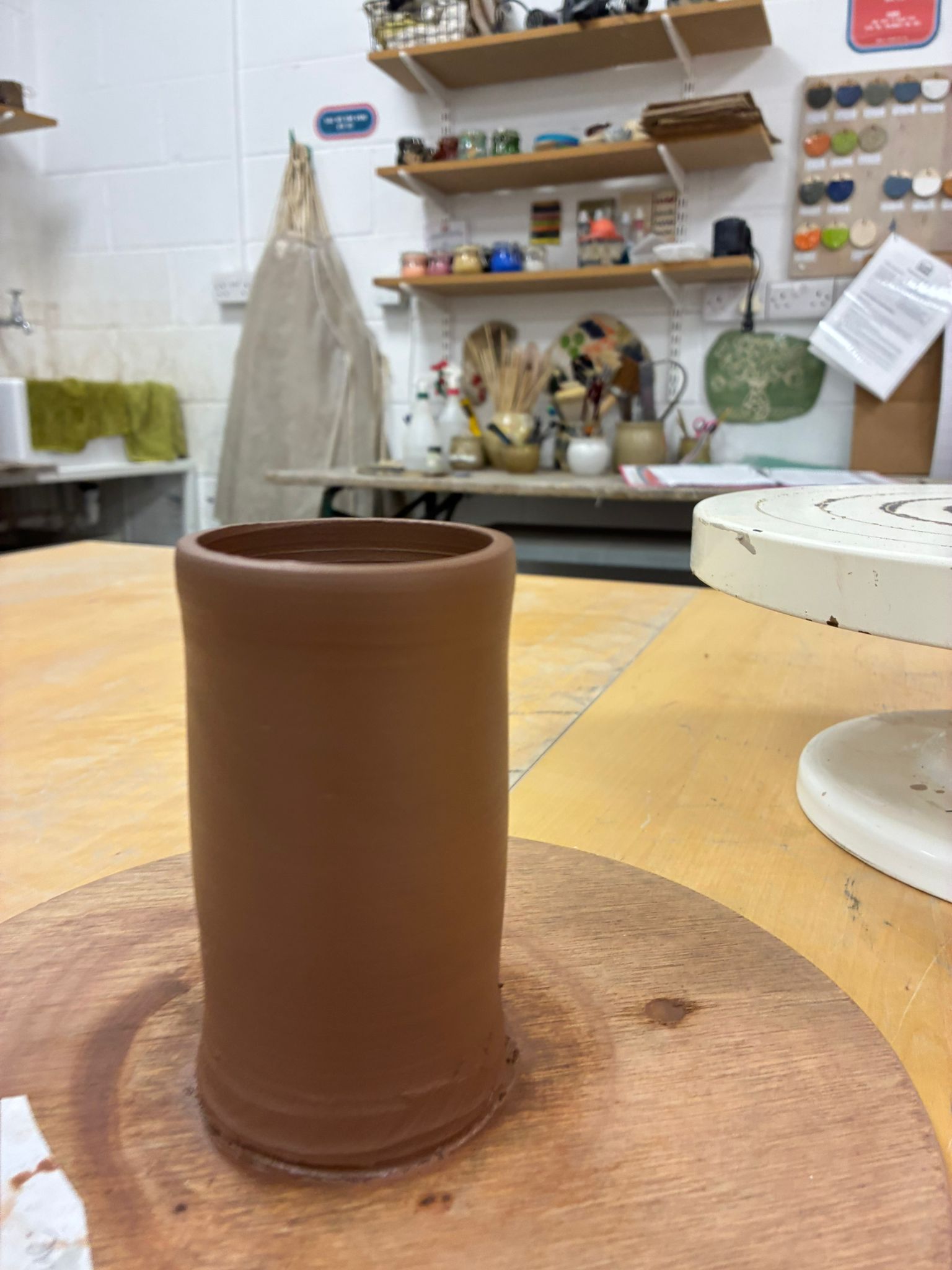 A tall cylindrical stoneware vessel, freshly thrown, resting on a bat in the studio with glaze samples visible on shelves behind