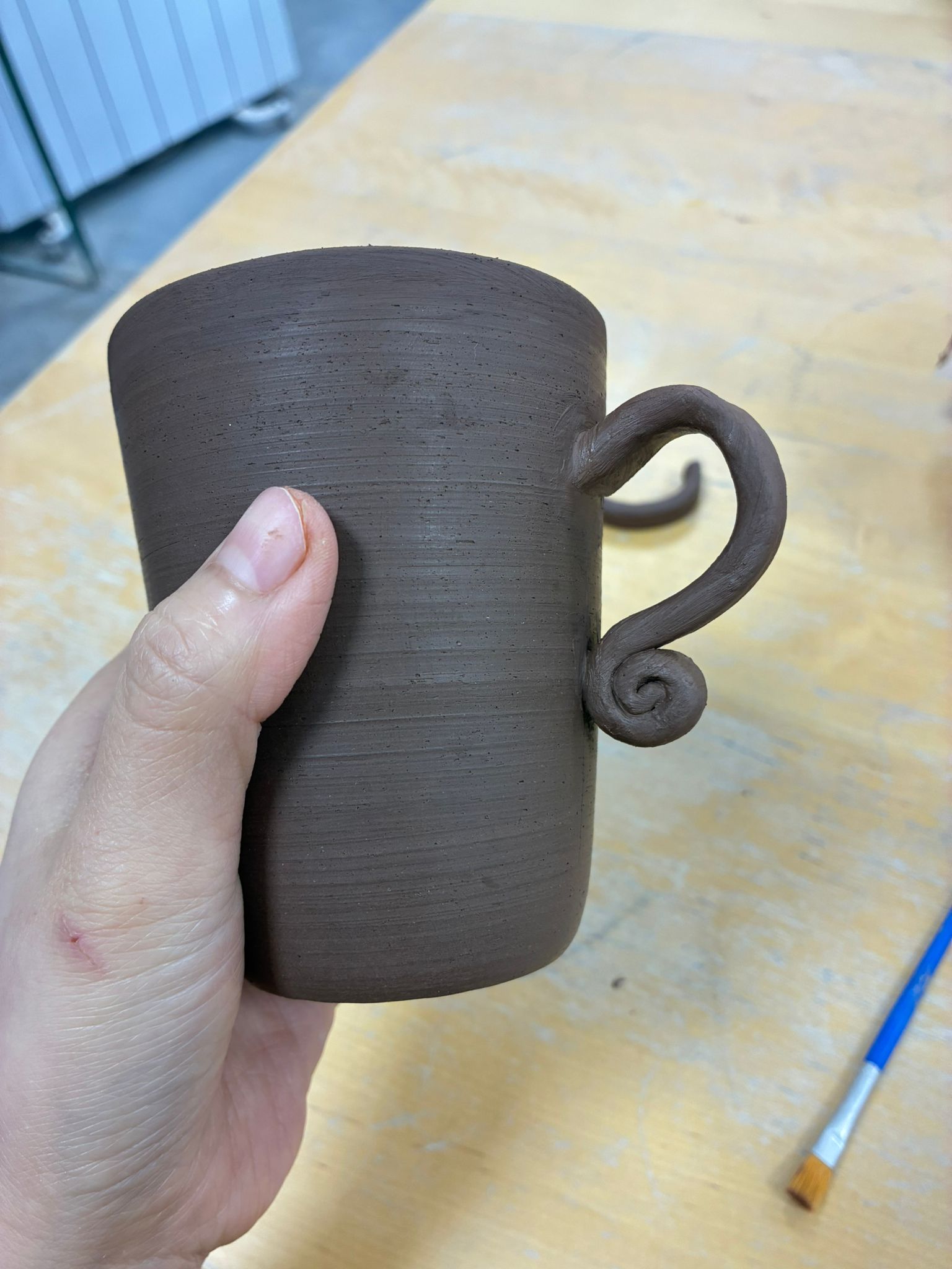 A hand holding a dark stoneware mug with a decorative curled scroll handle, raw and unfired on a studio workbench