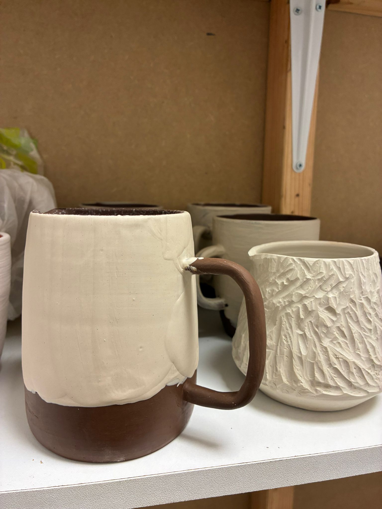 Two stoneware mugs on a studio shelf — one half-dipped in white slip over dark clay with a brown handle, the other with a deeply textured surface