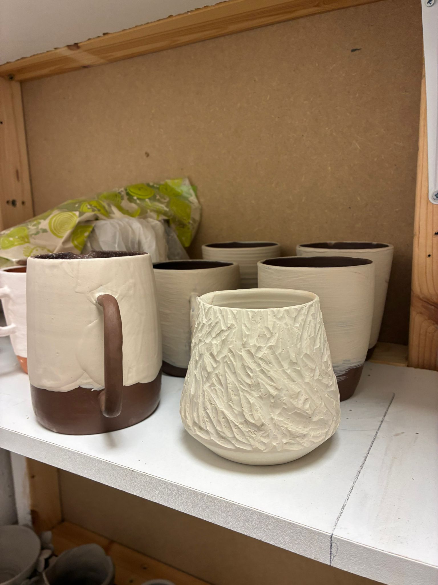 A group of works in progress on studio shelves — dipped mugs and a textured cup awaiting the kiln