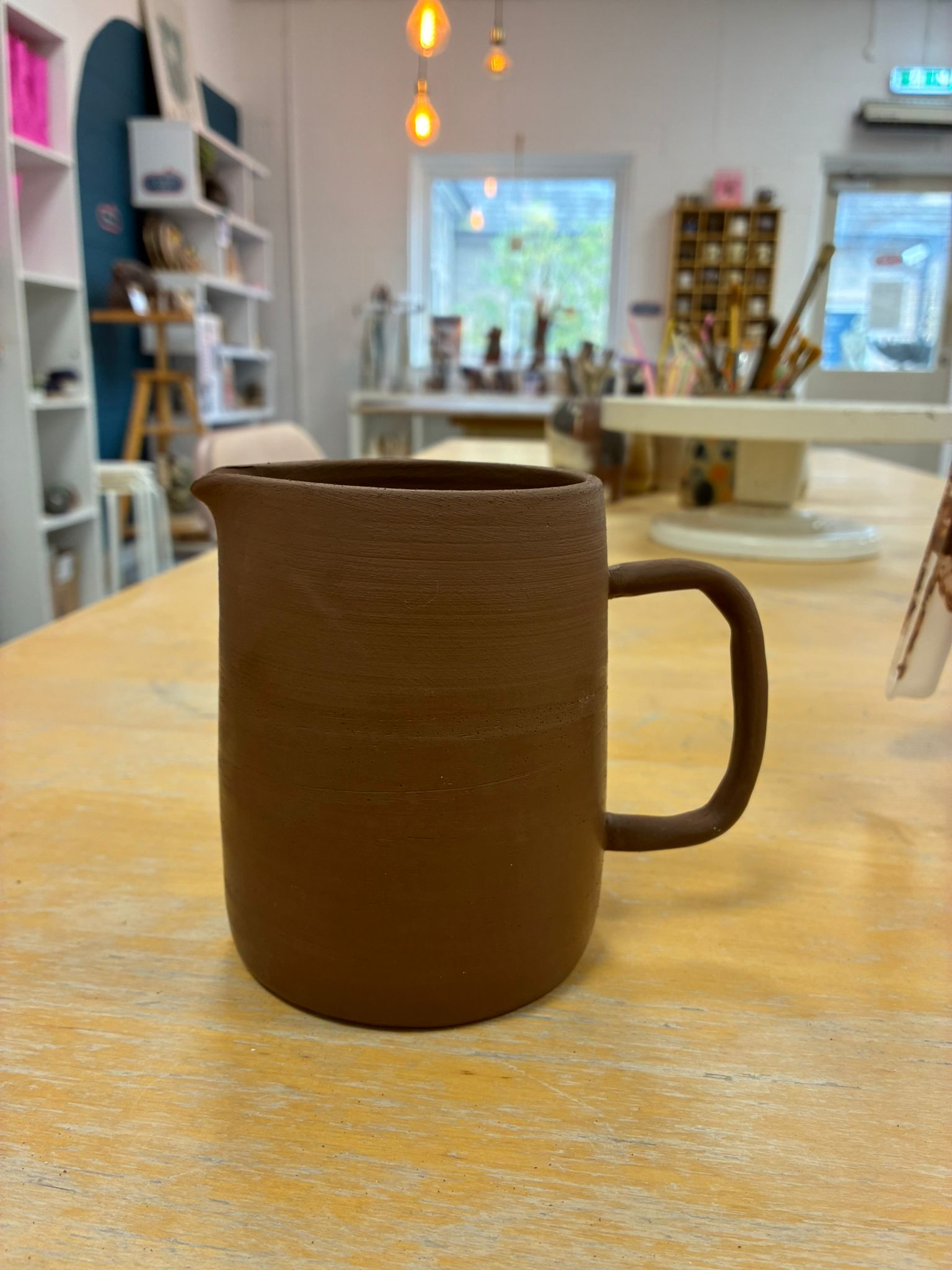 A smooth stoneware pitcher with a loop handle, raw and unfired, resting on a wooden studio workbench